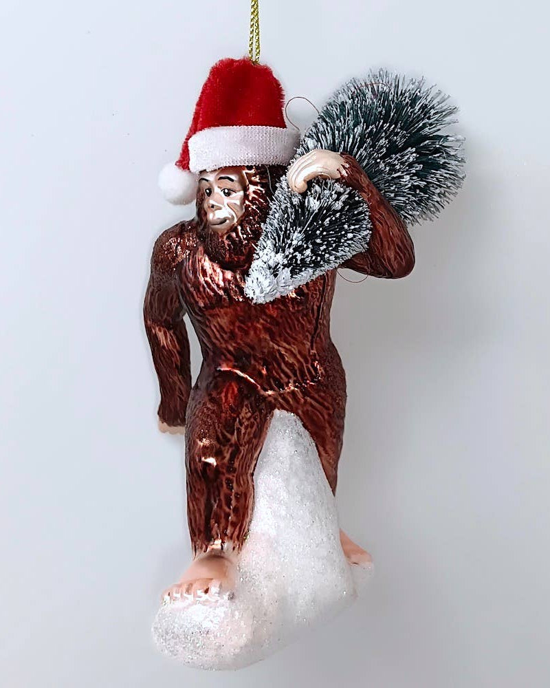 Christmas Cheer All Year Bigfoot glass ornament wearing Santa hat carrying christmas tree