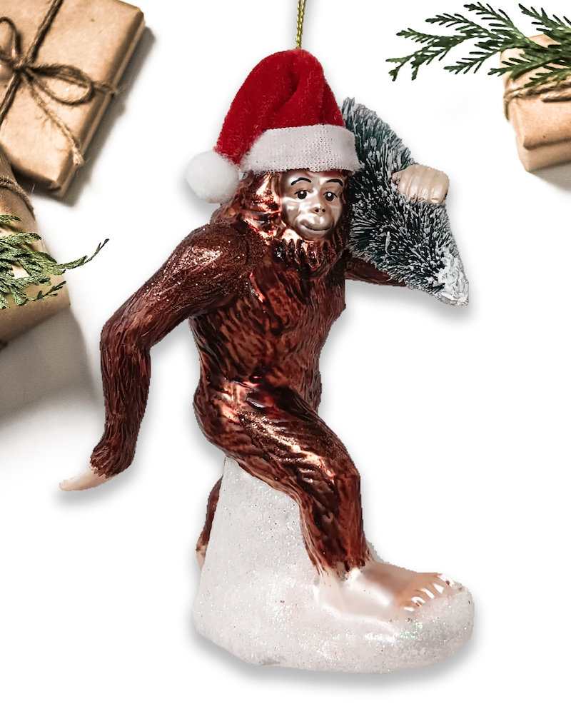 Christmas Cheer All Year Bigfoot glass ornament wearing Santa hat with packages