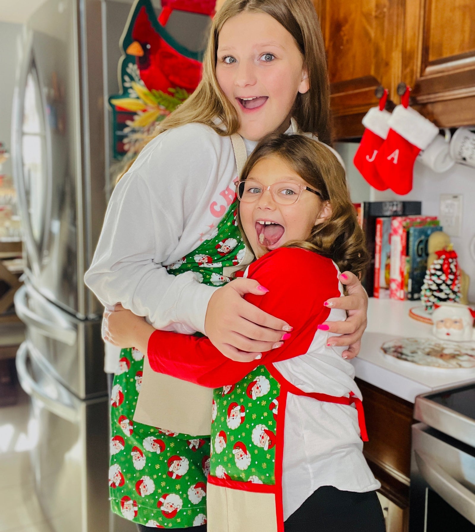 Two children in festive aprons hugging in a kitchen decorated for Christmas.