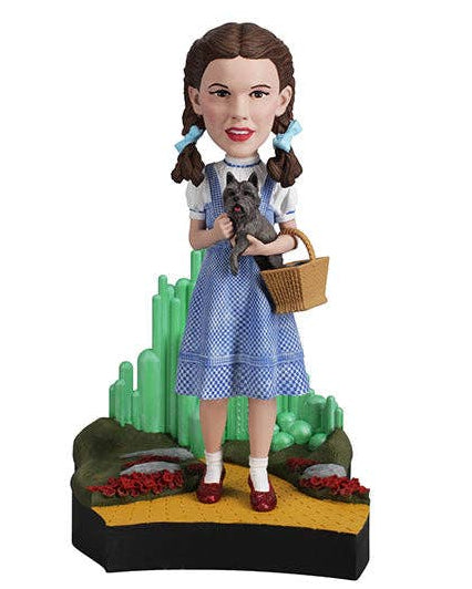 Christmas Cheer All Year A collectible figurine of Dorothy from The Wizard of Oz standing on a cactus hill with her cat, Toto, in her arms. She is wearing her signature blue and white gingham dress, white blouse, red shoes, and holds a brown wicker basket.