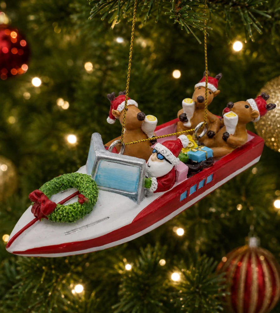 Christmas Cheer All Year Christmas ornament shaped like Santa Claus and his reindeer riding in a red and white speedboat, hanging from a gold string on a decorated Christmas tree with warm lights.