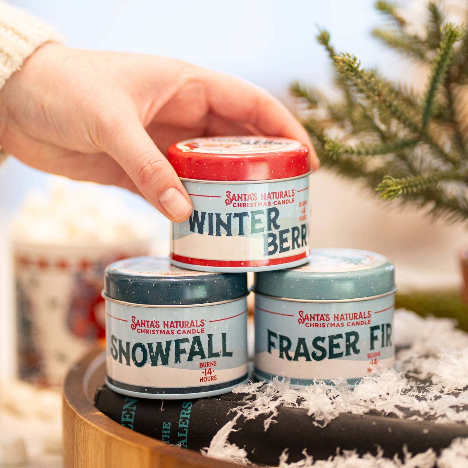 Christmas Cheer All Year Three small Santa's Naturals candles labeled 'Winter Berry', 'Snowfall', and 'Fraser Fir' on a wooden stand with a Christmas tree in the background.