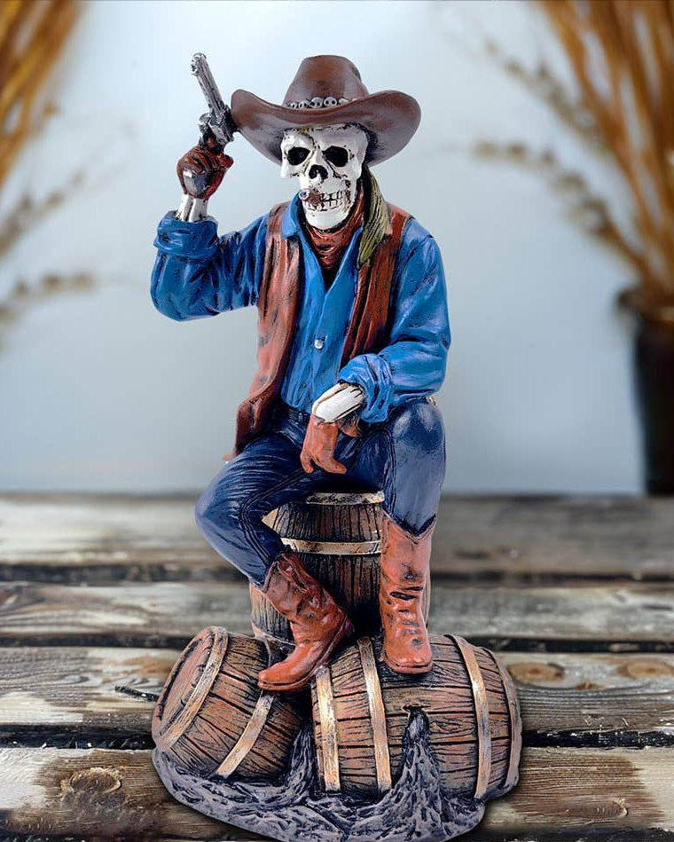 Christmas Cheer All Year handpainted figurine of a Western skeleton gunslinger skeleton wearing a cowboy hat, blue shirt, and brown vest, sitting on a barrel, holding a revolver, with details on the holster and boots.