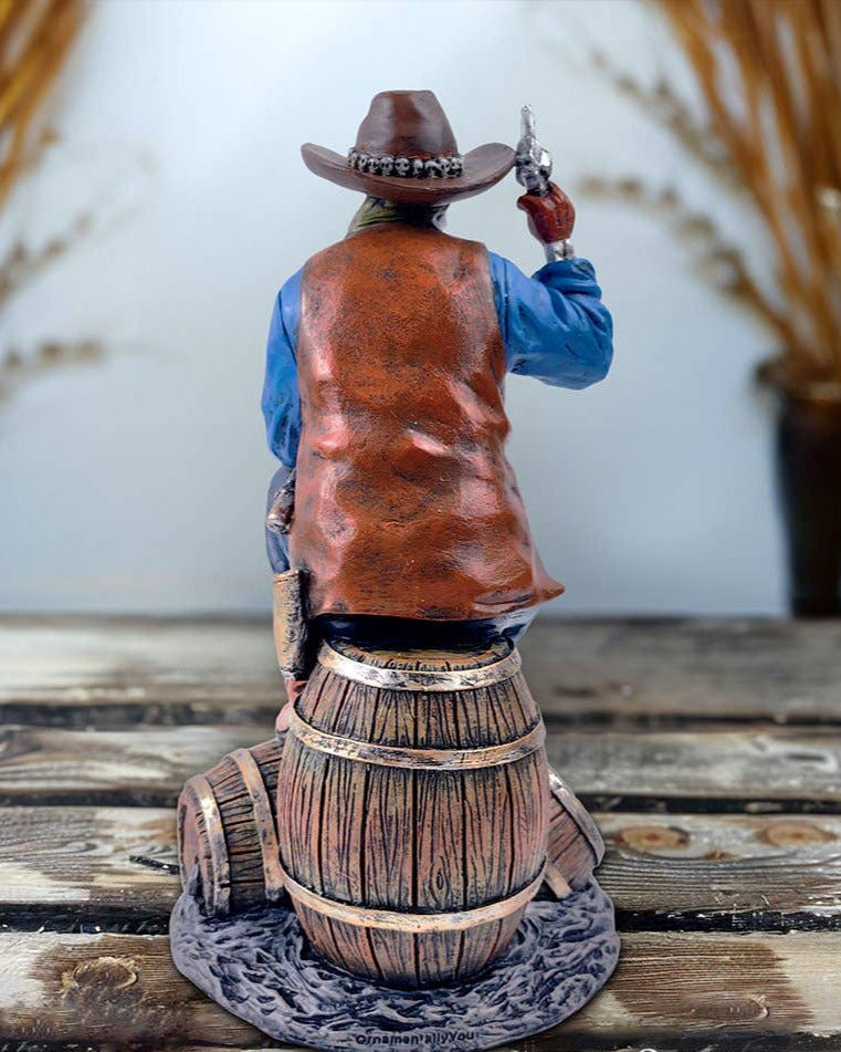 Christmas Cheer All Year handpainted figurine of a Western skeleton gunslinger skeleton wearing a cowboy hat, blue shirt, and brown vest, sitting on a barrel, holding a revolver, with details on the holster and boots.