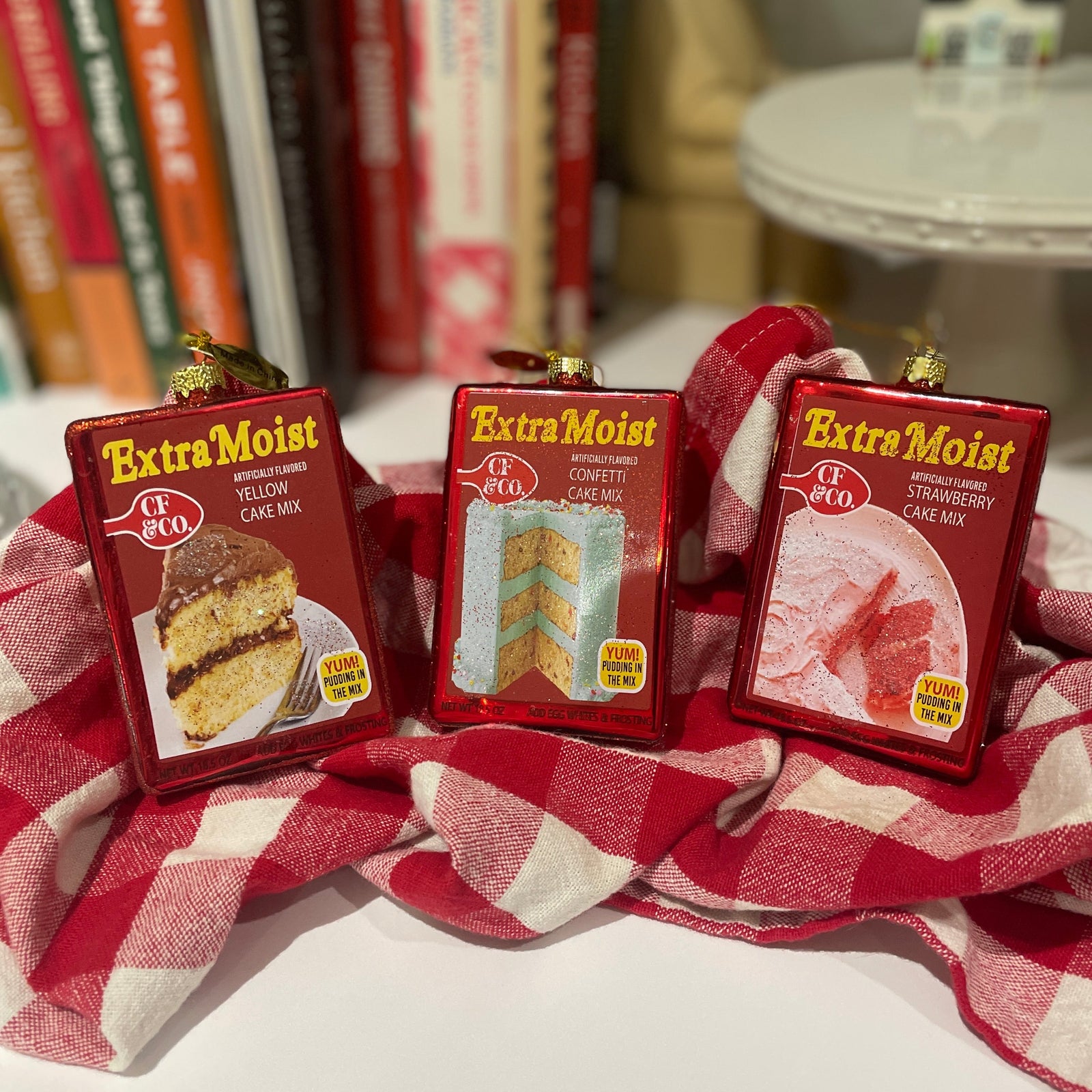 Christmas Cheer All Year Three boxes of 'Extra Moist' Betty Crocker cake mix ornaments by Cody Foster on a red and white checkered cloth 
