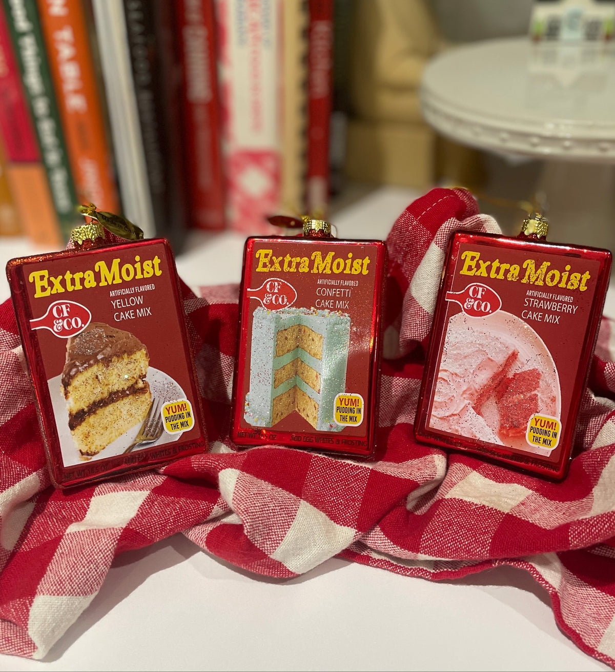 Christmas Cheer All Year Three boxes of 'Extra Moist' Betty Crocker cake mix ornaments by Cody Foster on a red and white checkered cloth 
