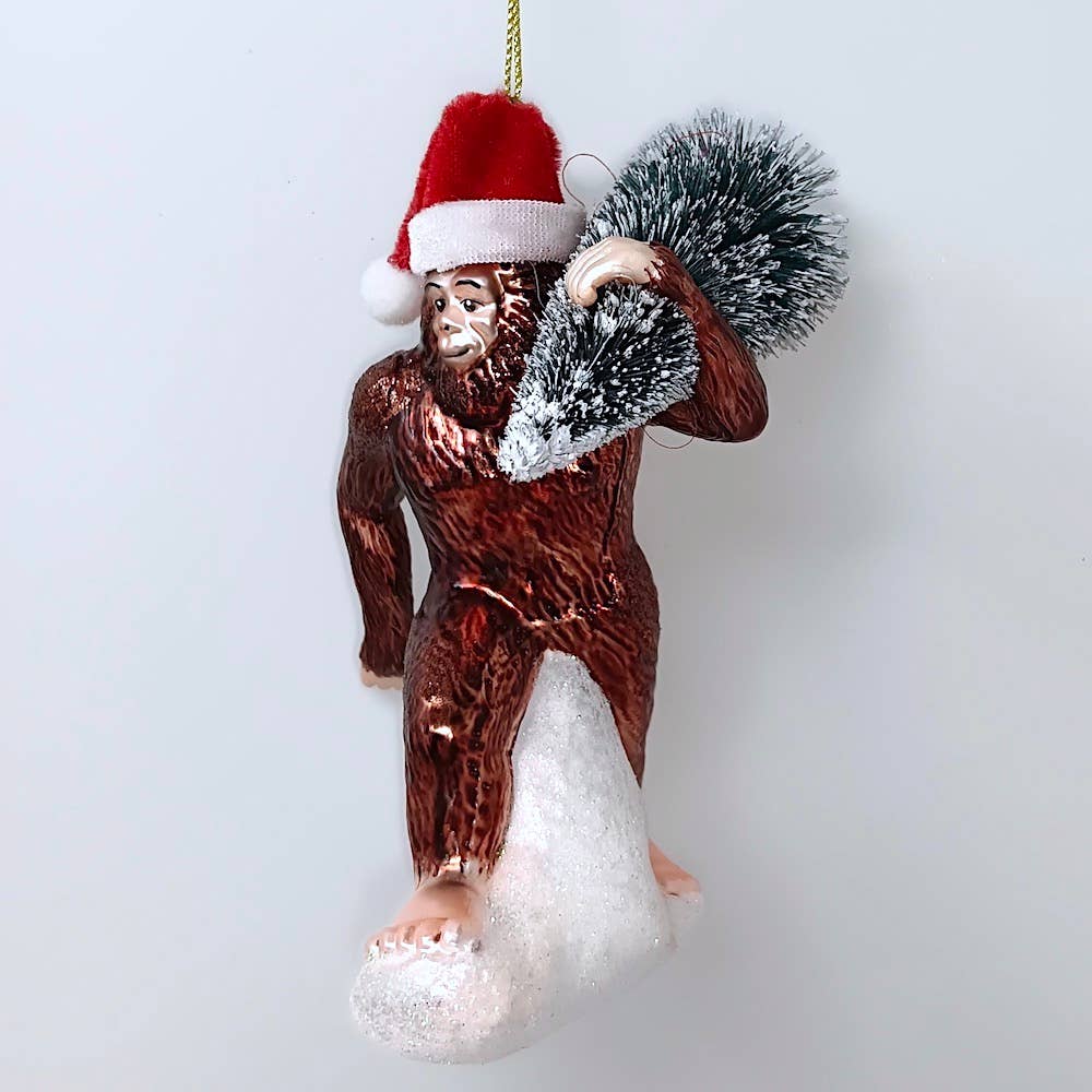 Christmas Cheer All Year Bigfoot glass ornament wearing Santa hat carrying christmas tree