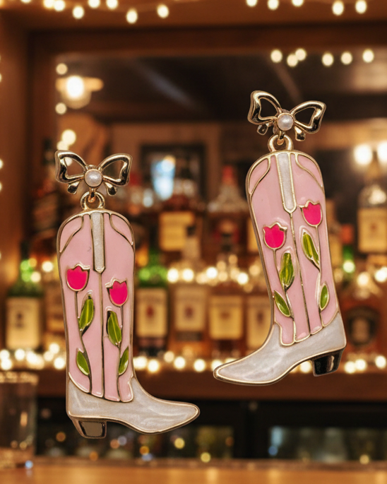 Christmas Cheer All Year dangling cowboy boot earrings with a pink and white faux-pearl finish, decorated with pink flowers and green leaves, and topped with gold bow details with pearl accents.