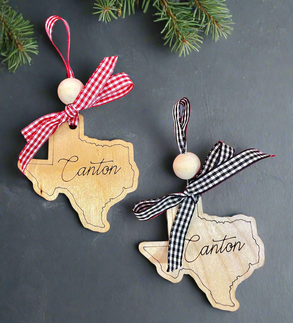 Two wooden ornaments in the shape of Texas with 'Canton' engraved on them, one with a red checkered ribbon and one with a black checkered ribbon Christmas Cheer All Year
