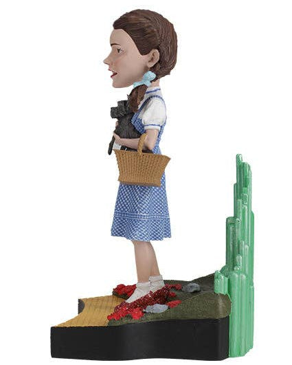 Christmas Cheer All Year A collectible figurine of Dorothy from The Wizard of Oz standing on a cactus hill with her cat, Toto, in her arms. She is wearing her signature blue and white gingham dress, white blouse, red shoes, and holds a brown wicker basket.