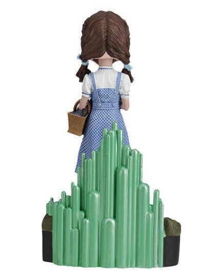Christmas Cheer All Year A collectible figurine of Dorothy from The Wizard of Oz, shown from behind wearing her blue and white gingham dress and white blouse, holding a brown basket. The figure stands on a green crystal mountain base against a solid white background.