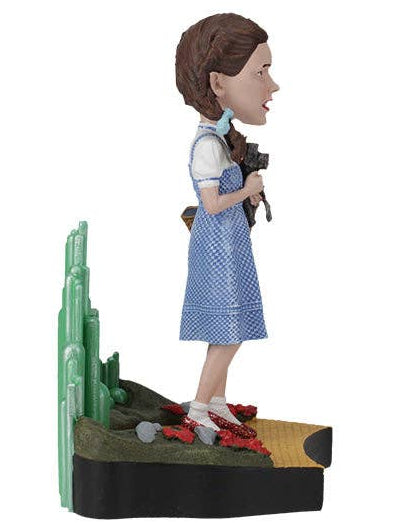 Christmas Cheer All Year collectible figurine of Dorothy from The Wizard of Oz standing in a field, wearing her blue and white polka-dot dress, white blouse, brown hair, and red shoes, posed next to green cacti on a black and tan base against a white background.