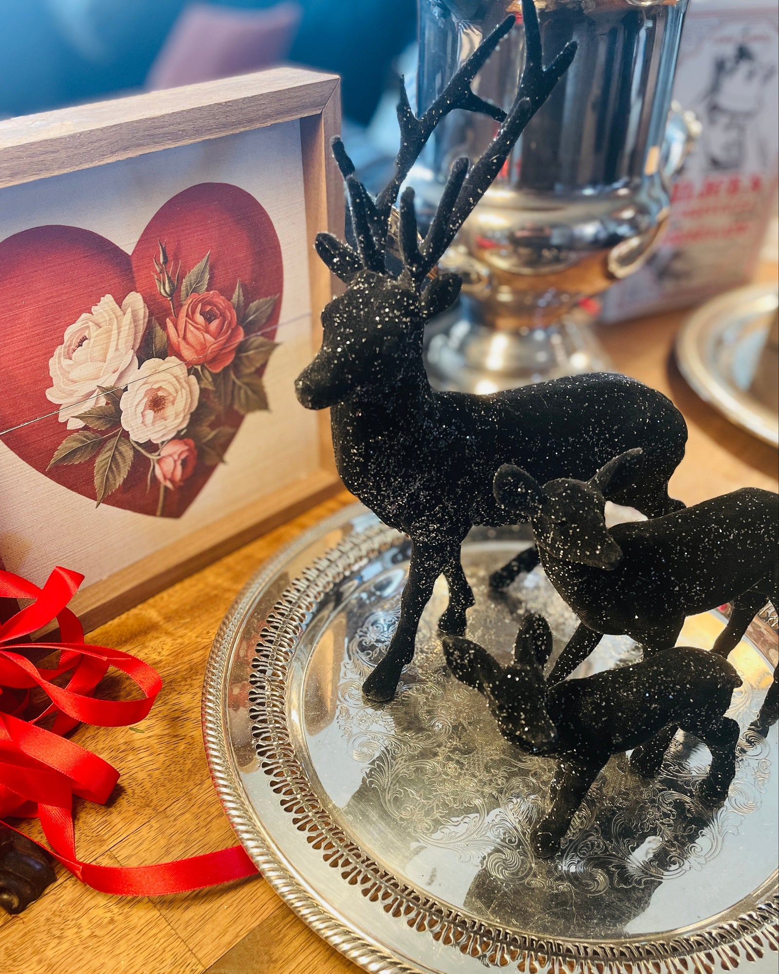 Christmas Cheer All Year A decorative black glittery stag and two fawn figurine set displayed on a round silver tray, with a red heart floral picture frame and red ribbon nearby on a wooden table.