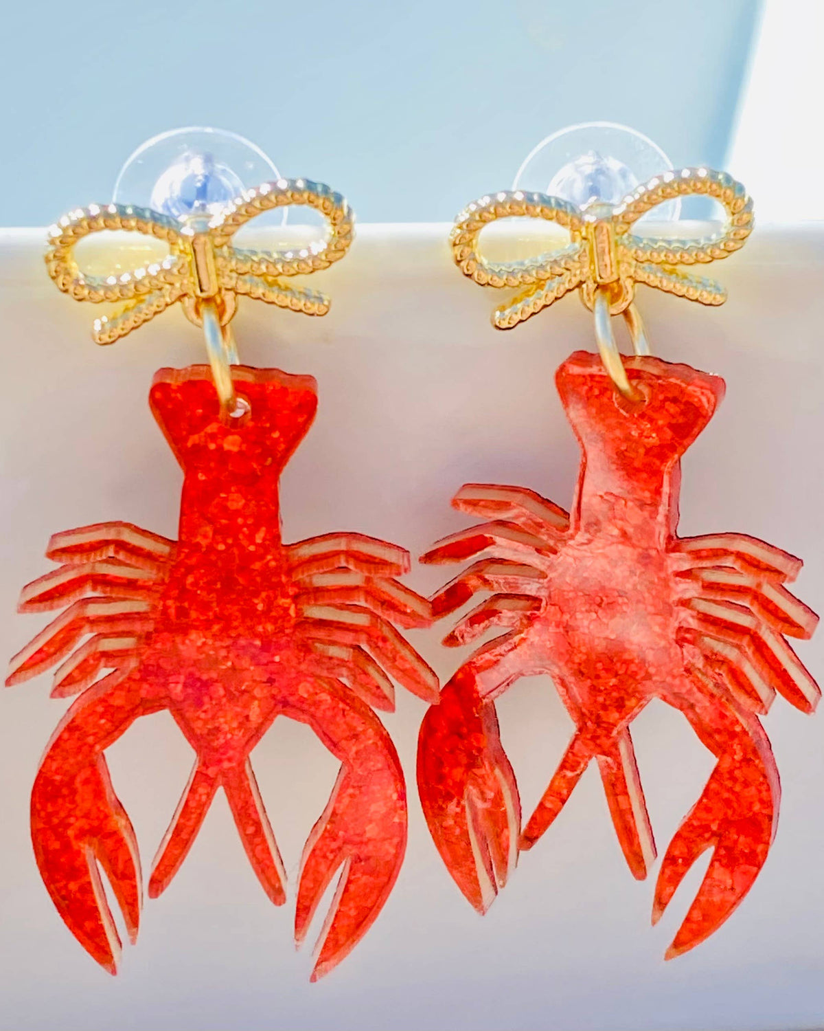 Red lobster-shaped earrings with gold bows on a light blue background