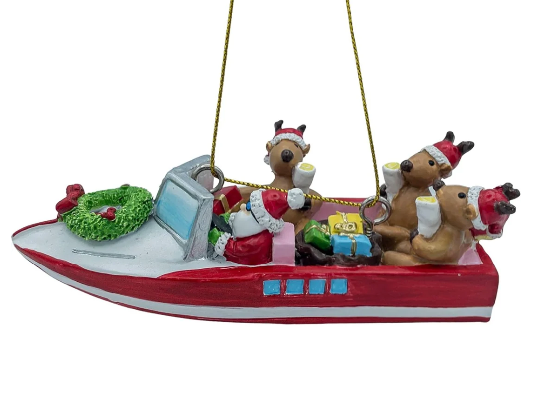 A Christmas ornament shaped like a red and white speedboat carrying Santa Claus, reindeer, gifts, and a green wreath, with gold hanging strings, shown on a plain white background.