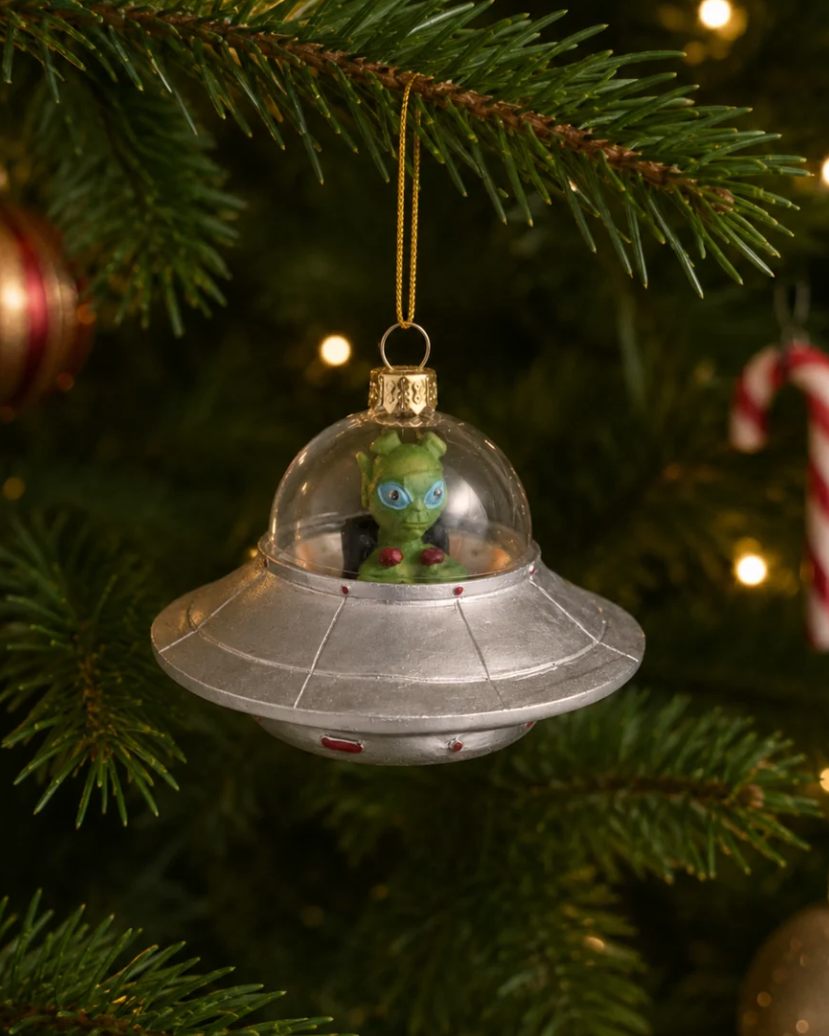 Christmas Cheer All Year silver domed alien spacecraft with green alien inside Christmas ornament hanging from a pine tree branch. The alien is inside a clear domed silver UFO spacecraft with a gold hanging loop