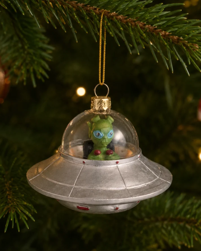 Christmas Cheer All Year silver domed alien spacecraft with green alien inside Christmas ornament hanging from a pine tree branch. The alien is inside a clear domed silver UFO spacecraft with a gold hanging loop