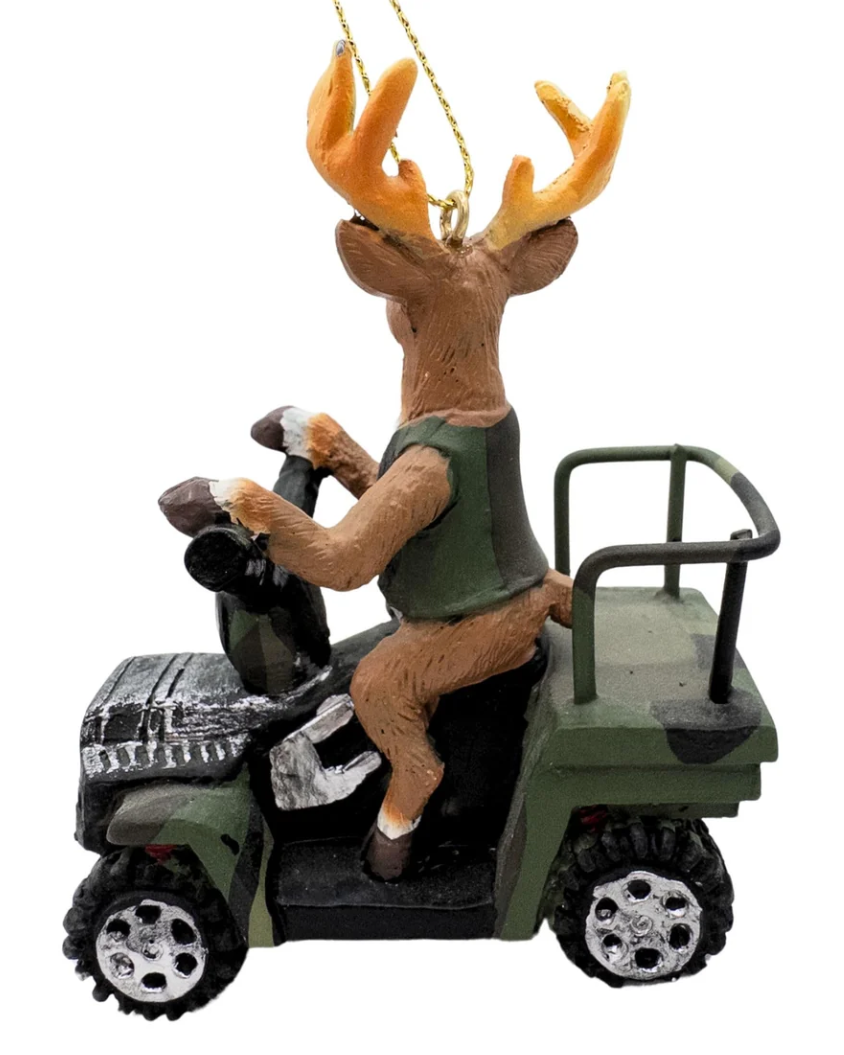 Christmas Cheer All Year A deer ornament driving an ATV 4 wheeler, hanging from a pine tree with warm string lights in the background.