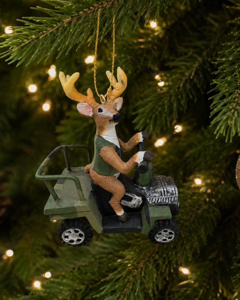 Christmas Cheer All Year A deer ornament driving an ATV 4 wheeler, hanging from a pine tree with warm string lights in the background.
