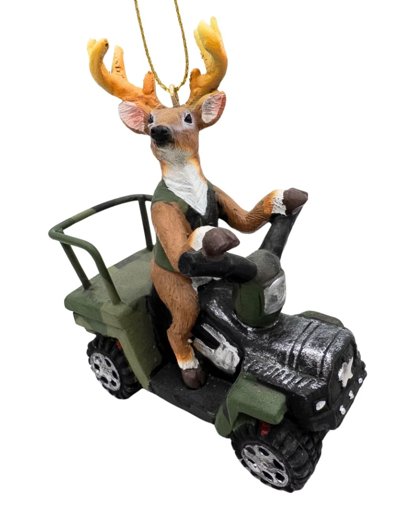 Christmas Cheer All Year A deer ornament driving an ATV 4 wheeler, hanging from a pine tree with warm string lights in the background.