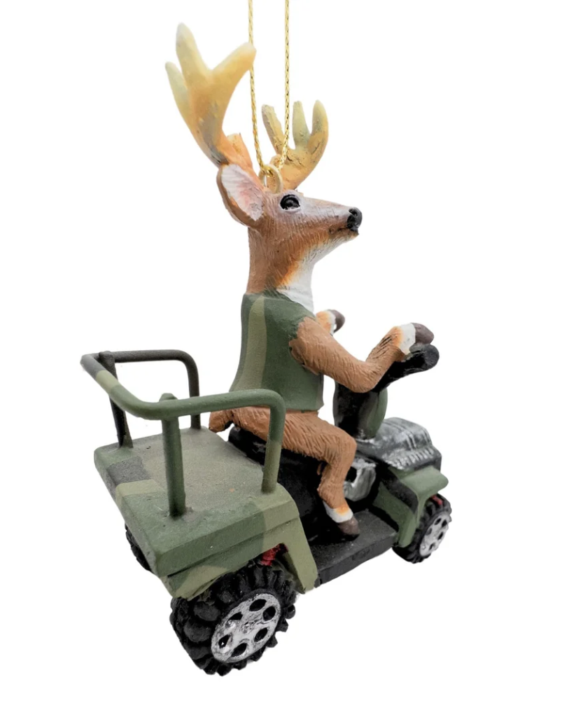 Christmas Cheer All Year A deer ornament driving an ATV 4 wheeler, hanging from a pine tree with warm string lights in the background.