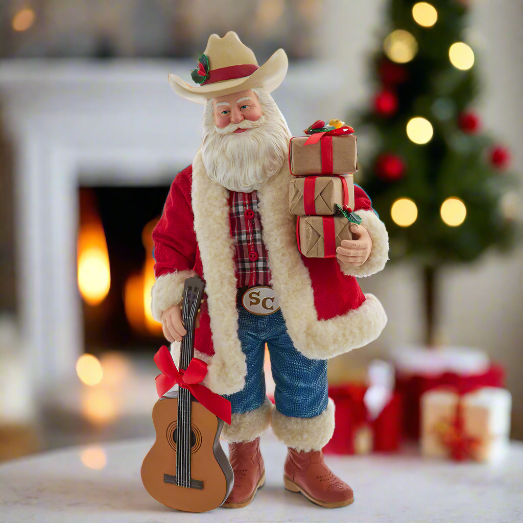 Christmas Cheer All Year Decorative Santa Claus Cowboy figure FA0217 holding a guitar and gift boxes in a festive setting with a fireplace and Christmas tree.
