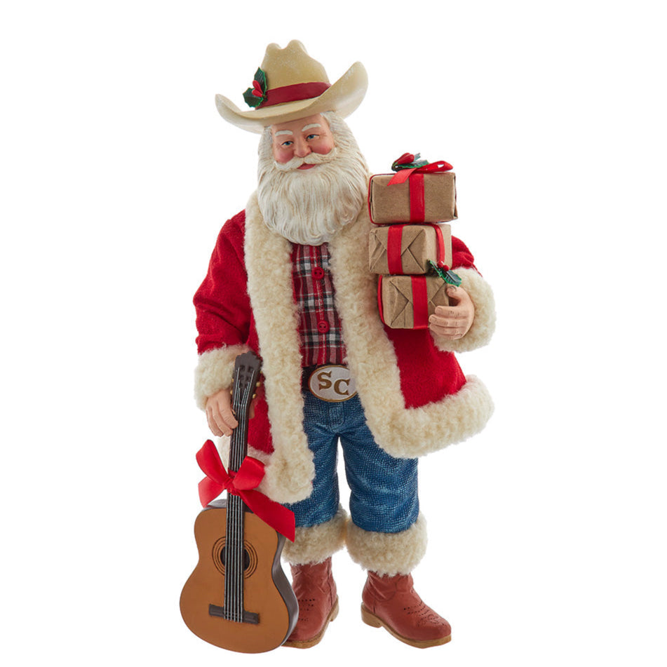 Christmas Cheer All Year Decorative Santa Claus Cowboy figure FA0217 holding a guitar and gift boxes in a festive setting with a fireplace and Christmas tree.